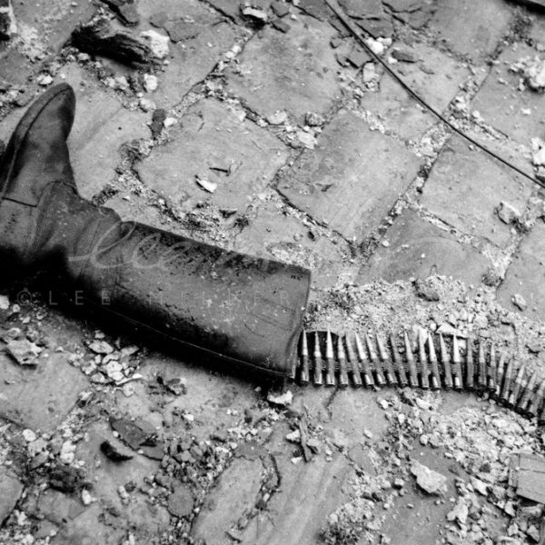 Black and white. Close up of soldier's boot with bullet belt coming out of it, amongst debris on the ground.