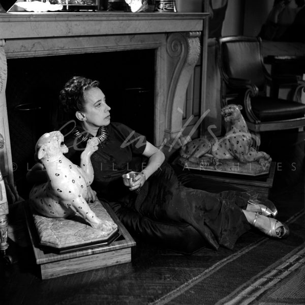 Black and white. Schiaparelli, seated on floor by fireplace with leopard sculptures.