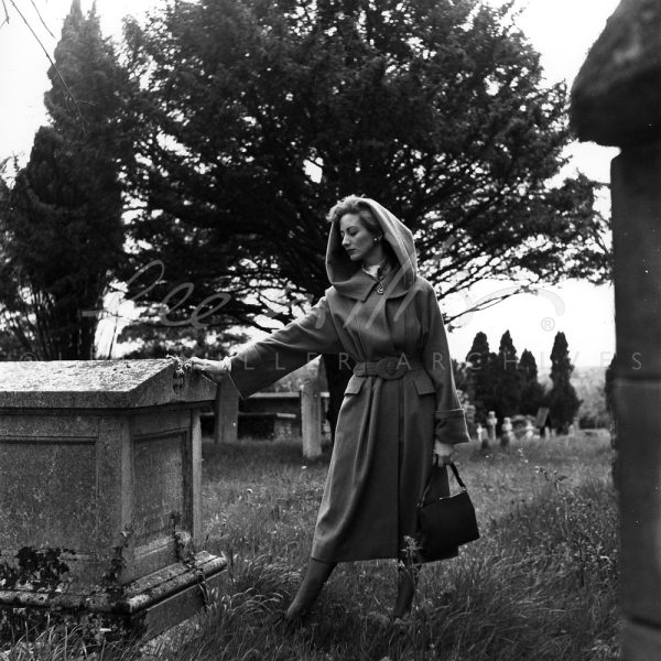 Black and white. Model in hooded coat leaning on a tomb in the churchyard.