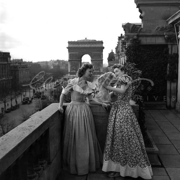 Black and white. Two models in long gowns pose on a roof top near the Arc de Triomphe.
