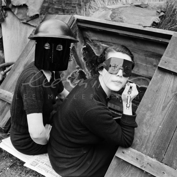 Black and white. Two women wearing firemasks sitting in the entrance to an air raid shelter. Their backs are towards the viewer and they are turning towards the camera. Their legs are inside the shelter.