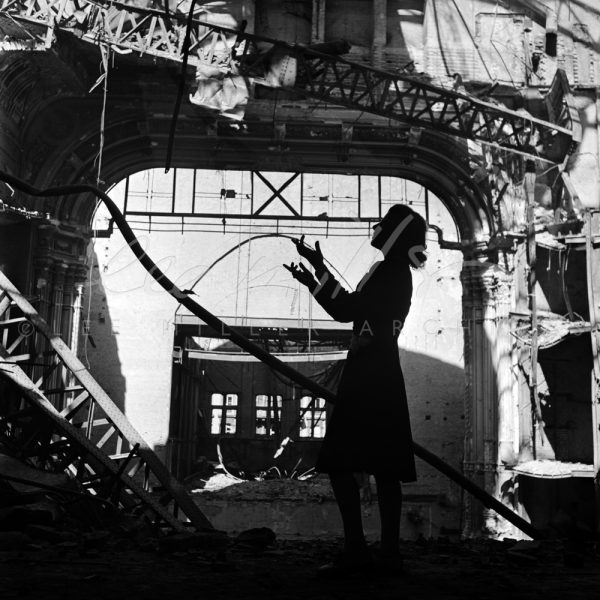 Black and white. Silhouetted figure with arms raised standing in bomb damaged building.