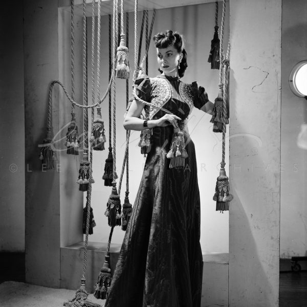 Black and white. Fashion model in long gown posing with tasselled ropes in studio.