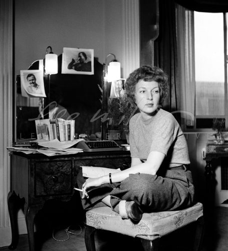 Black and white. Martha Gellhorn sits cross-legged by her dressing table.