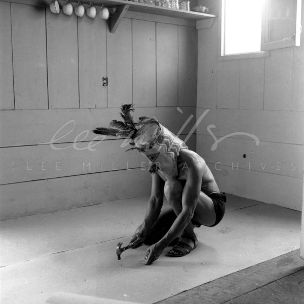 Black and white. In a newly built studio Max Ernst crouches to nail flooring in place. His head obscured by a large mask with feathers.