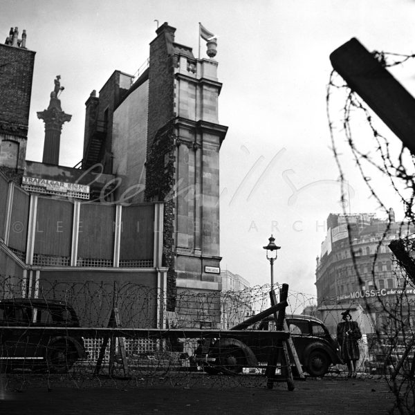 Black and white city scene during the Blitz. Nelson's Column can be seen through a gap in the buildings. A board advertises 'Trafalgar Signs'. To the right a view of buildings in the distance. In the foreground barbed wire defences, motor vehicles and a fashion model wearing a hat and fur coat.