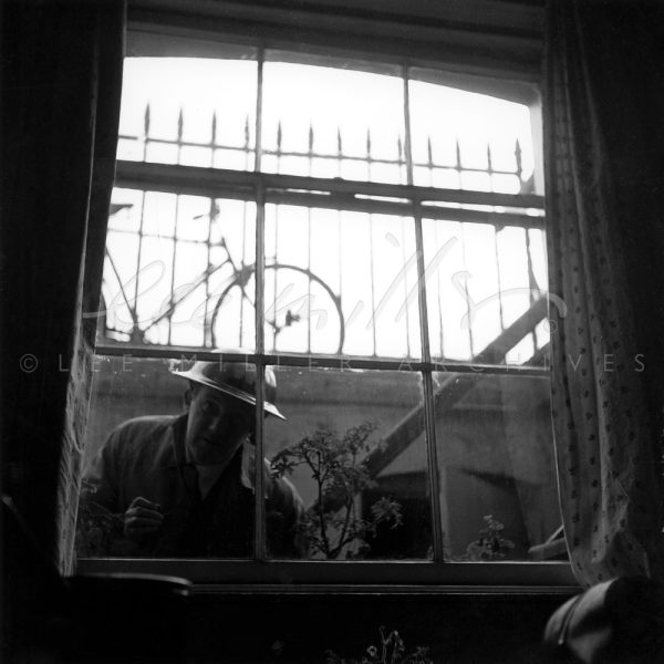 Black and white. View through window from basement interior. Air raid warden in tin hat looking in.
